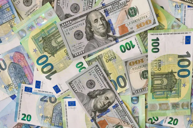 Billetes de $, € y otras divisas junto a una pantalla con tipos de cambio.