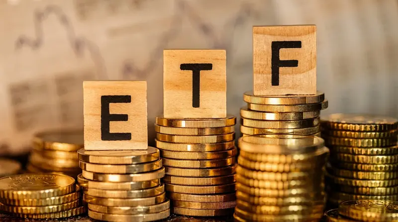 Las letras E, T y F en bloques de madera sobre pilas de monedas doradas, con un gráfico bursátil de fondo.