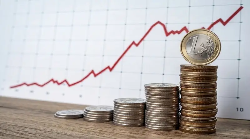 Pilas de monedas de € cada vez más altas, con una moneda de 1 € encima y un gráfico ascendente de fondo.