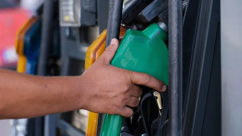 Una mano sujeta la pistola verde de un surtidor de gasolina, con otros surtidores desenfocados de fondo