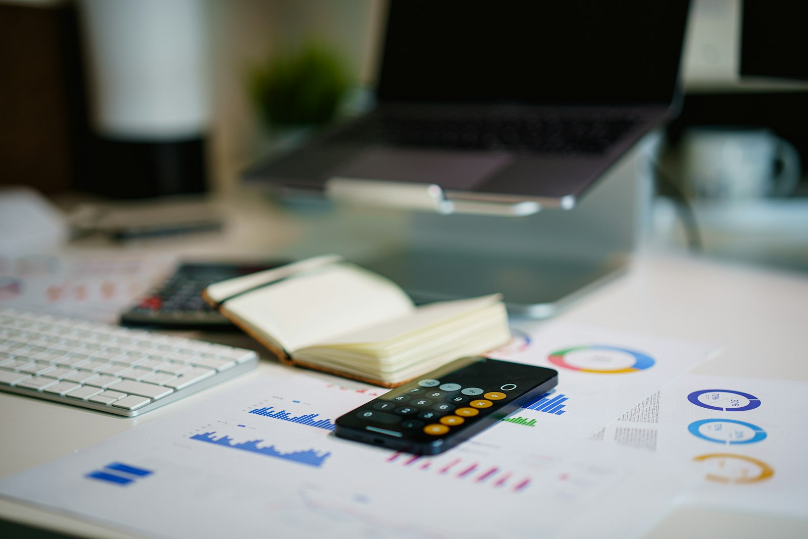 Gráficos financieros impresos sobre un escritorio, junto a un teléfono con calculadora, un cuaderno y un portátil.