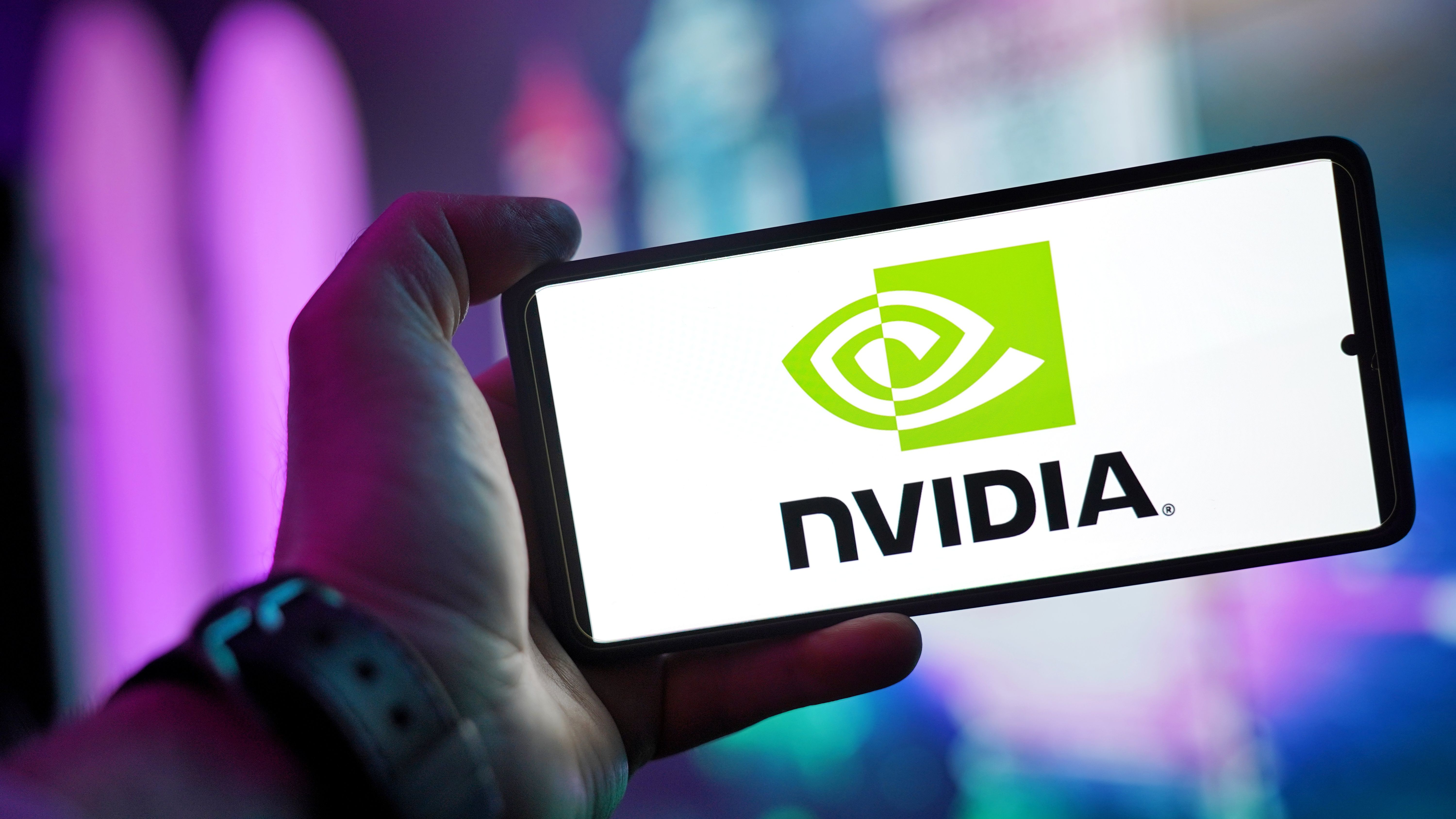 Una persona sostiene un teléfono que muestra el logo de Nvidia en la pantalla, con luces violeta y azul de fondo.