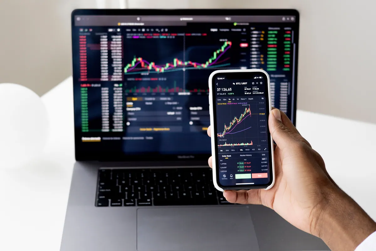 Gráfico de velas japonesas con medias móviles y osciladores técnicos en un teléfono, frente a una terminal de trading en un portátil.