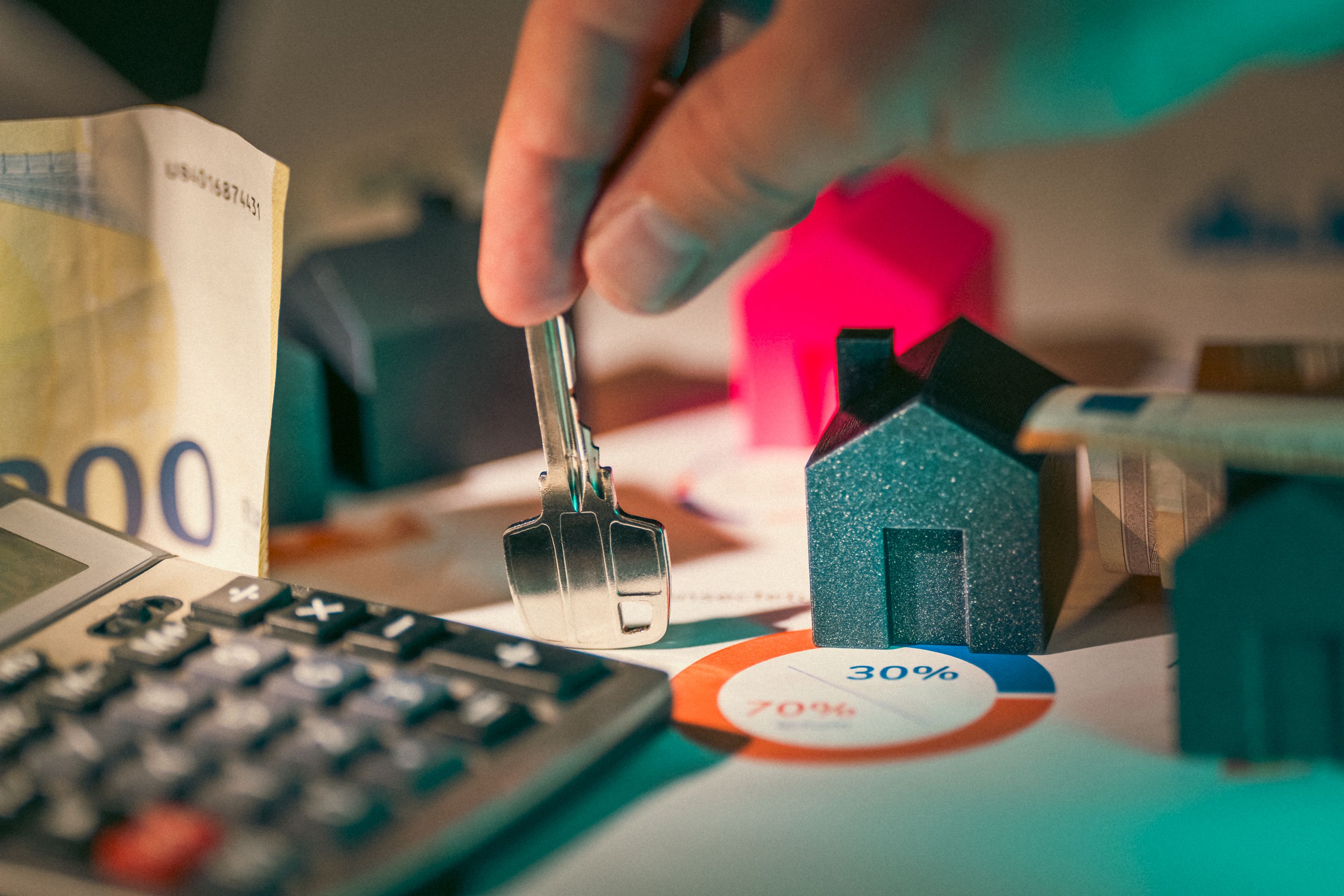 Una mano sostiene la llave de una vivienda junto a una calculadora, billetes de € y una casa miniatura, con un gráfico circular al 70%/30% de fondo.