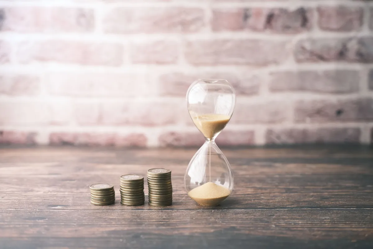 Tres pilas de monedas cada vez más altas junto a un reloj de arena, como metáfora del dinero que crece con el paso del tiempo.