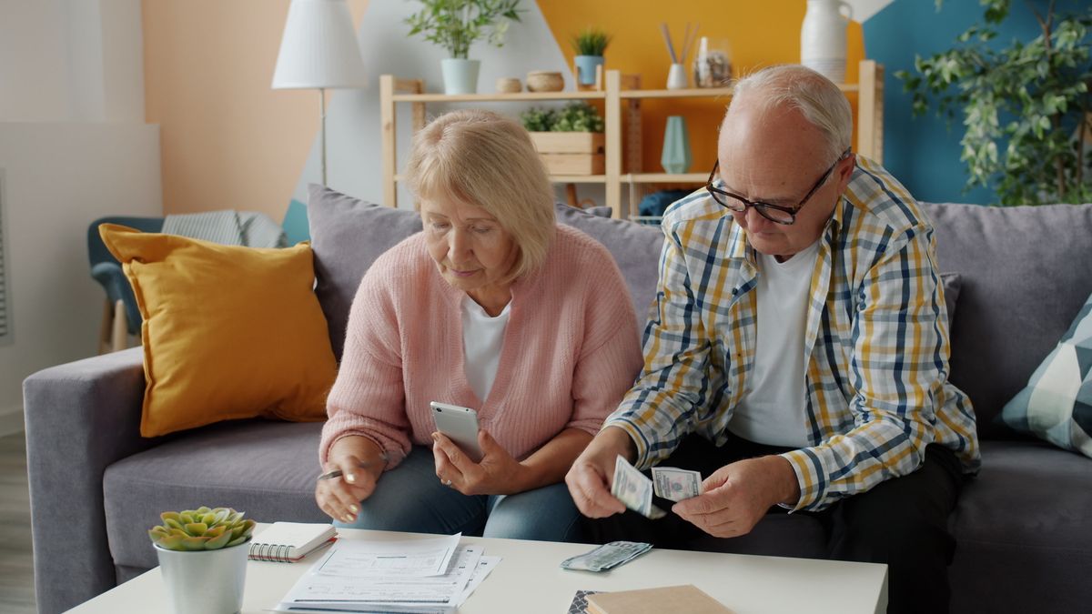 Una pareja de jubilados revisa sus cuentas en el sofá con un móvil, billetes y documentos.