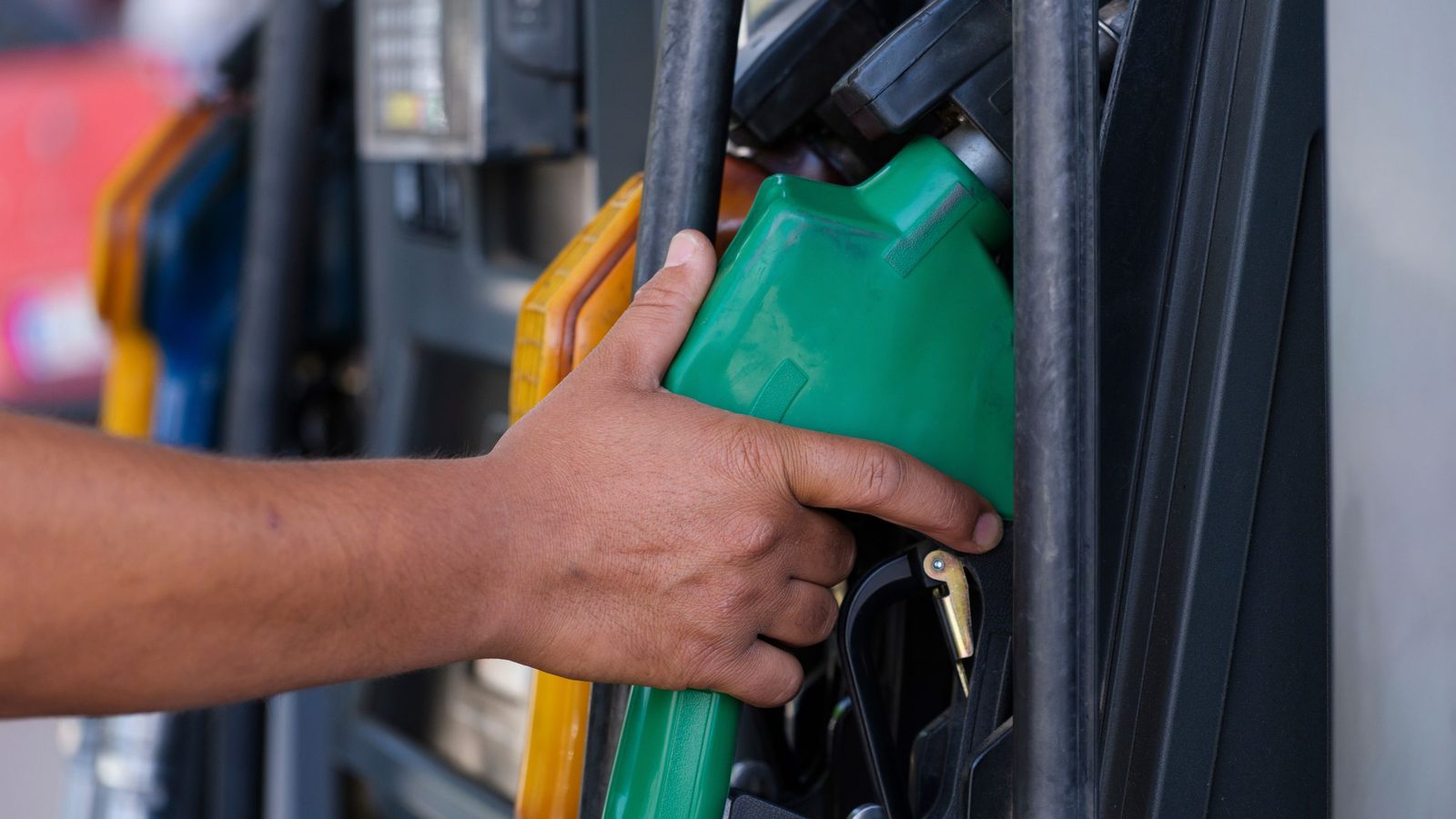 Una mano sujeta la pistola verde de un surtidor de gasolina, con otros surtidores desenfocados de fondo